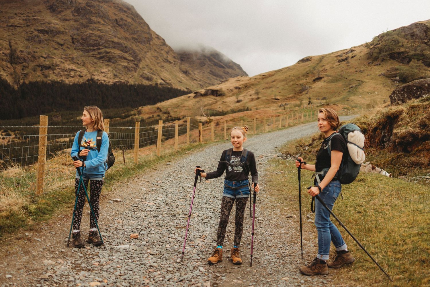 Three hikers with backpacks and poles on a mountain trail surrounded by rugged Scottish highlands.