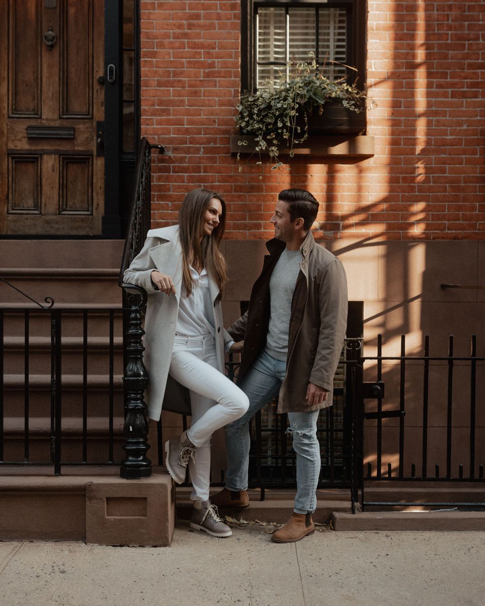 A stylish couple in casual attire posing against a sunlit brick wall and iron railing.