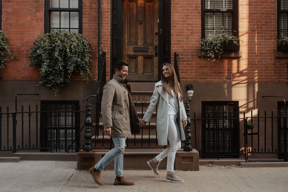 A couple in matching light coats walking together outside a classic brownstone building.