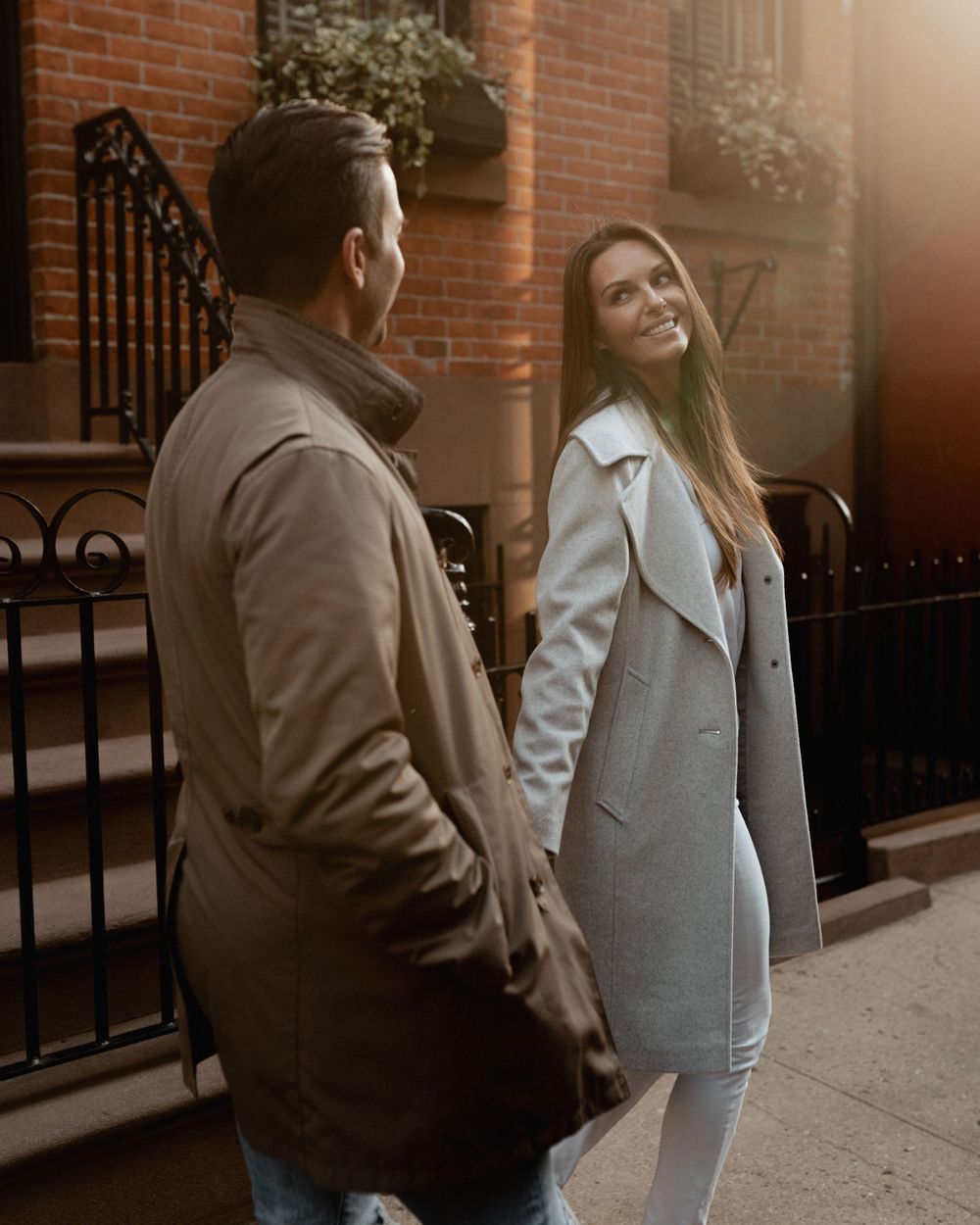 A couple sharing a playful moment in warm sunlight outside a brick townhouse.