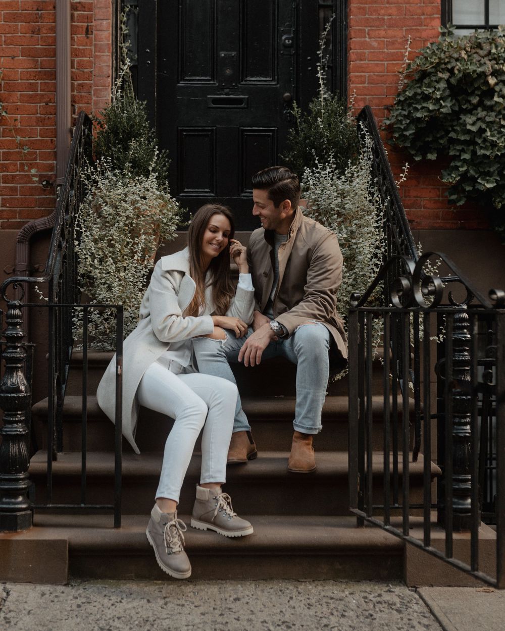 A couple sitting close together on brownstone steps with decorative plants framing the entrance.