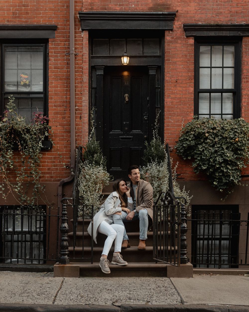 A couple sitting on the steps of a classic brick townhouse with black door and window frames.