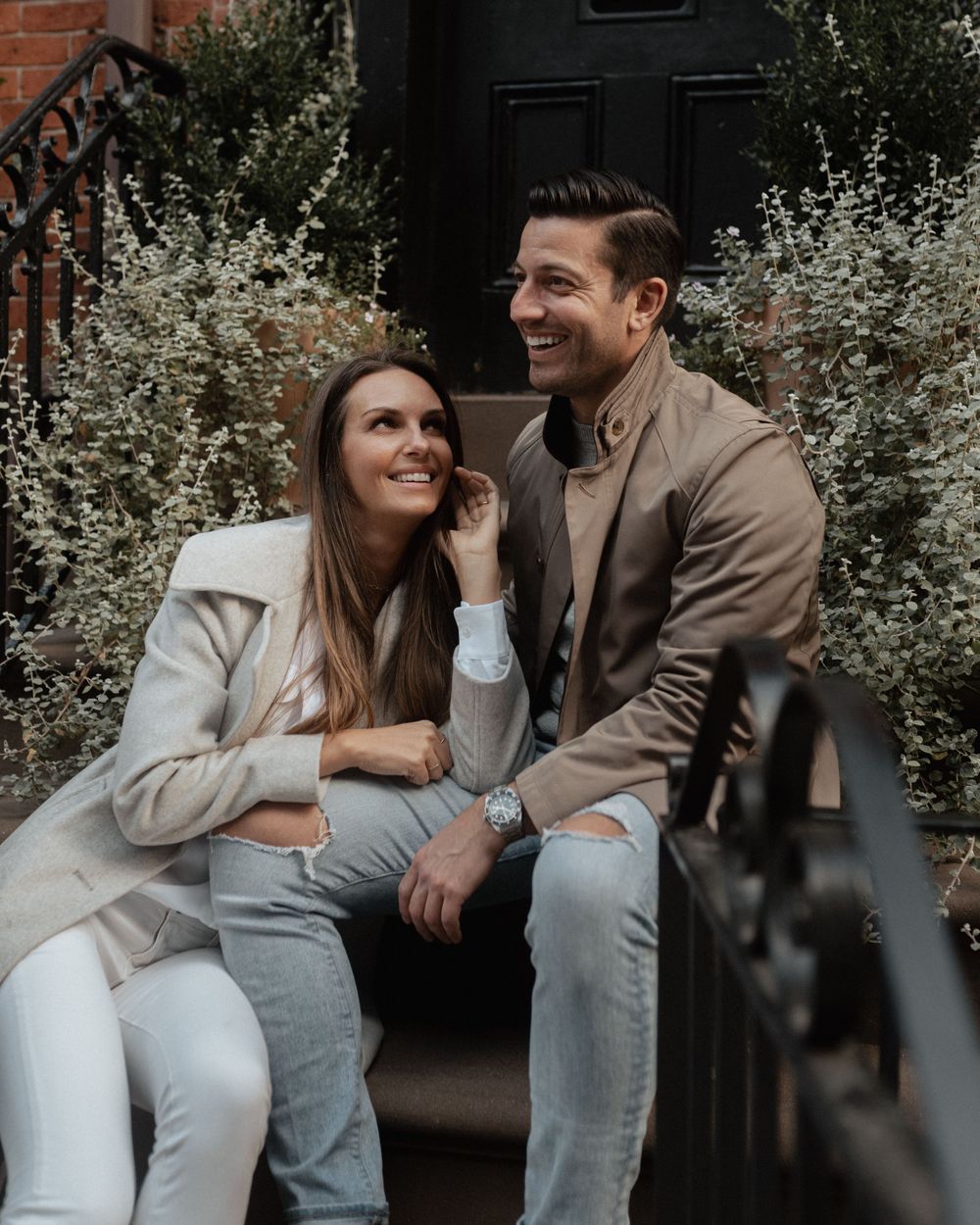 Couple in casual attire sharing intimate moments while sitting outdoors near flowering bushes.