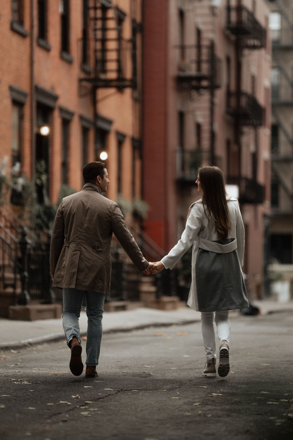 Couple walking hand in hand down a moody urban street with brownstone buildings.