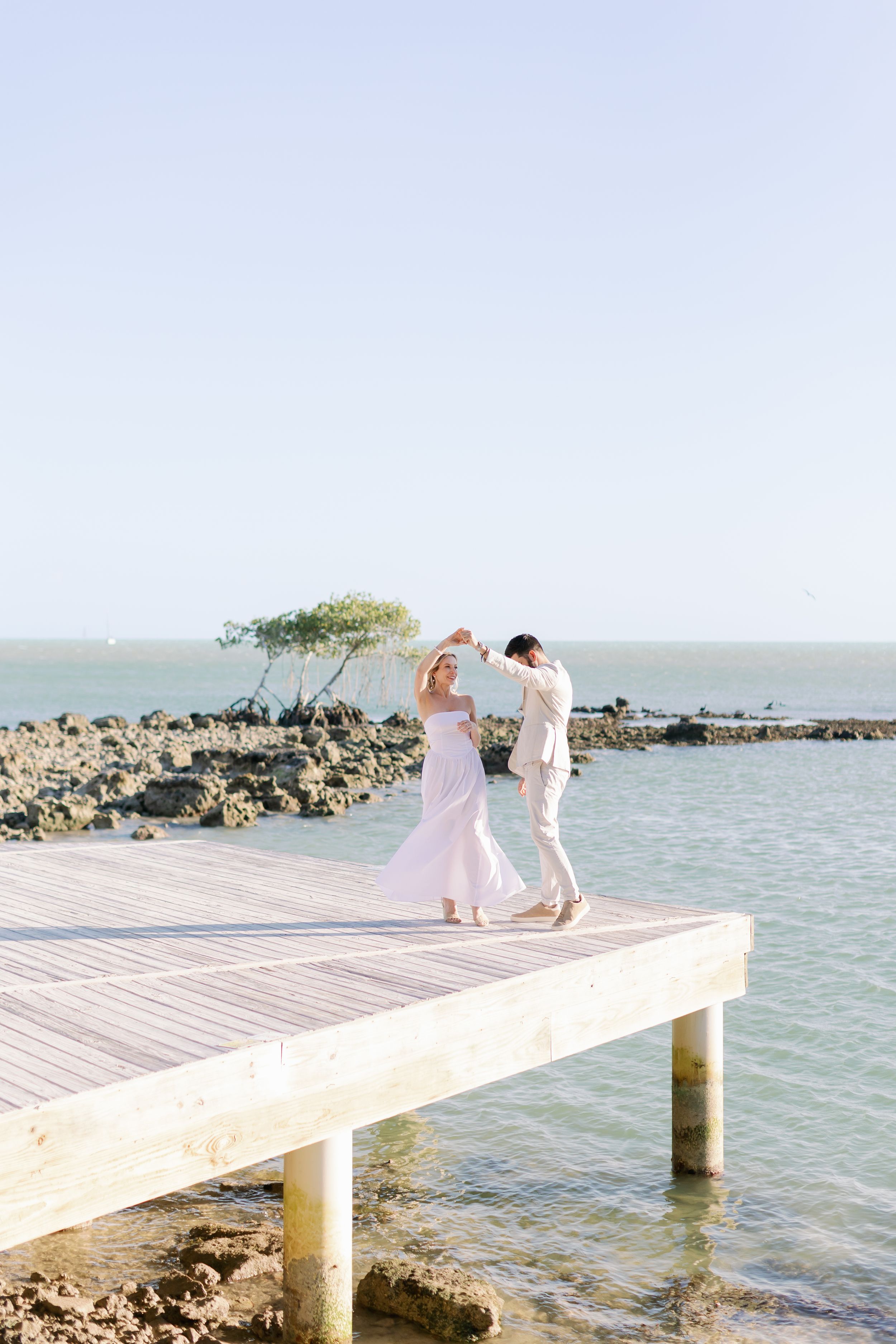 A couple stands together on a wooden pier overlooking tropical waters with rocky coastline in the background.