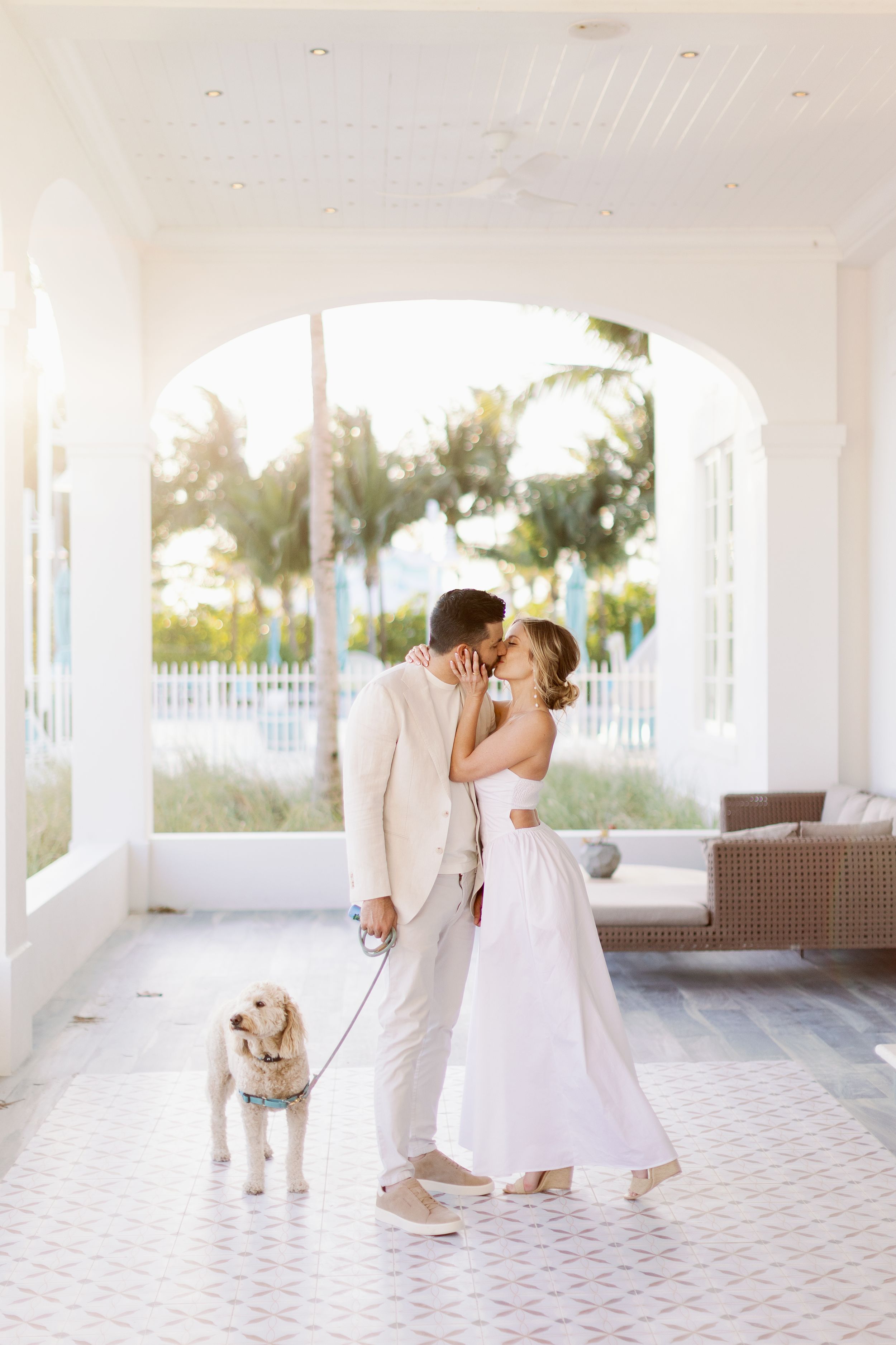 A couple in elegant white attire shares a romantic moment on a porch with their golden retriever puppy on a leash.