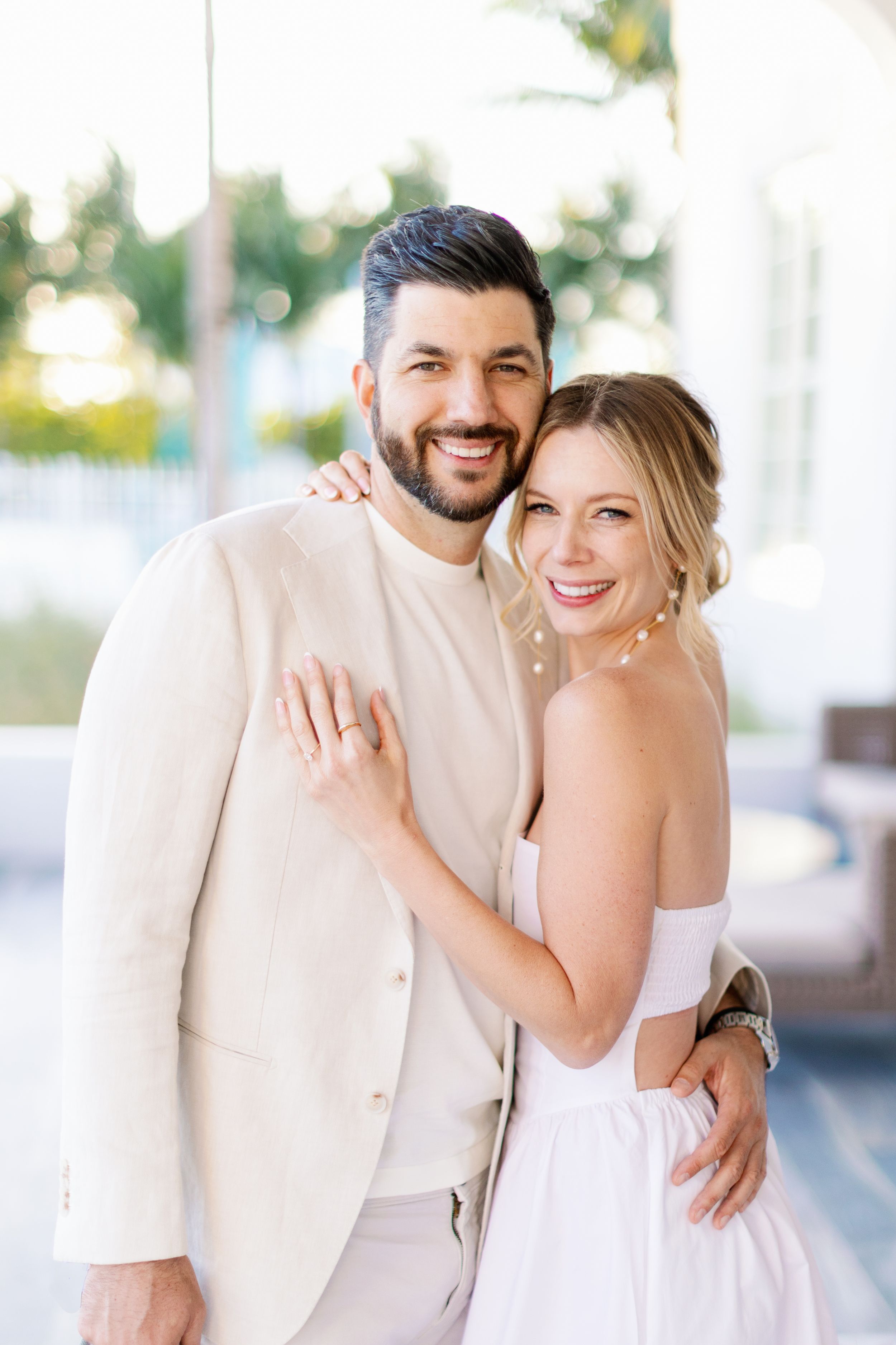 Happy couple in light-colored wedding attire embracing and smiling together outdoors on a sunny day.
