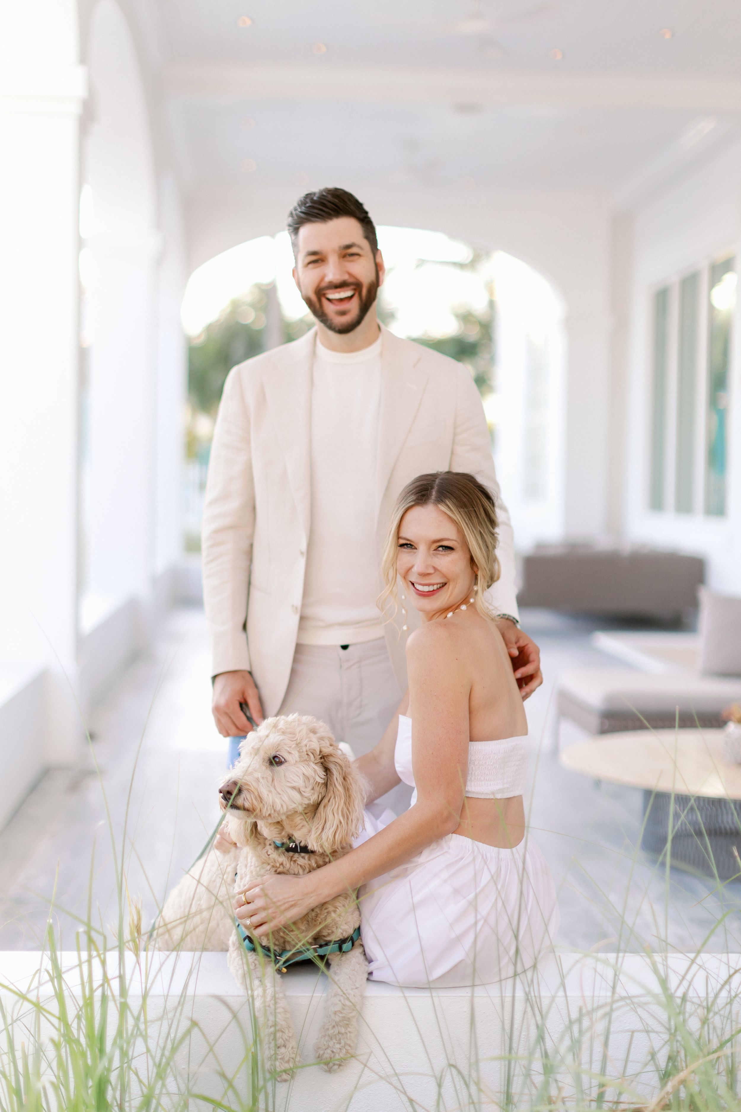 Couple poses on white porch with their fluffy white dog, wearing coordinated cream and white outfits.