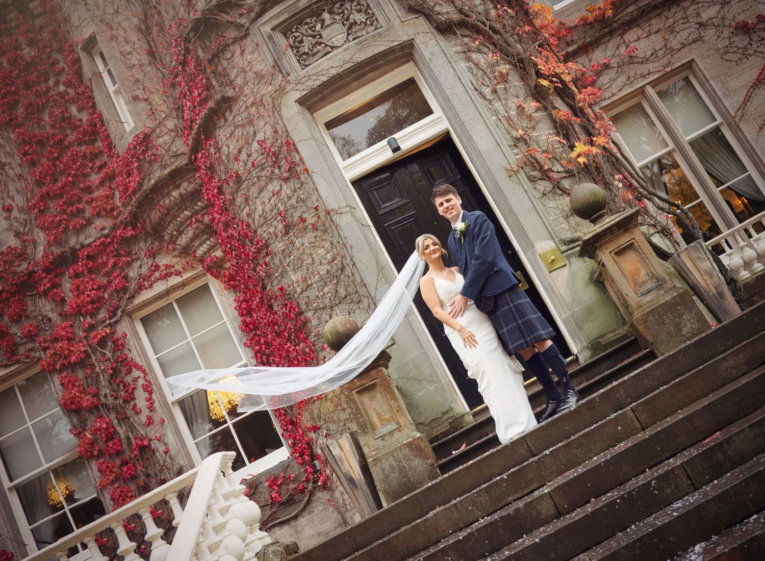 A bride and groom posing on their wedding day at Carlowrie castle. Veil is blowing in the wind and the husband arm protects his wife tenderl