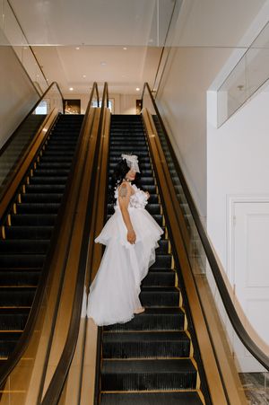 A person in a flowing white wedding gown ascends a modern escalator with gold railings in an elegant interior space.