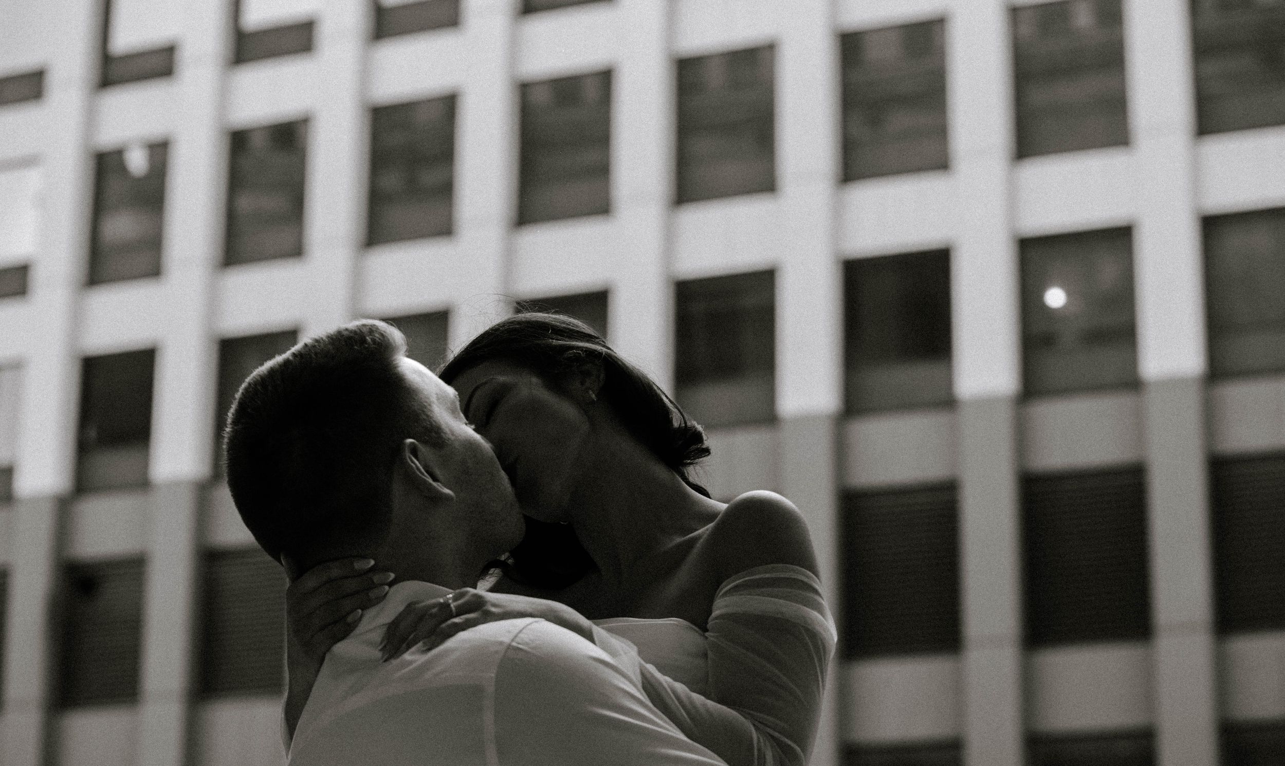 Two people share an intimate moment in front of an office building in black and white photography.