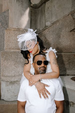 A couple in white attire embraces intimately, with one wearing a decorative white headpiece and sunglasses.
