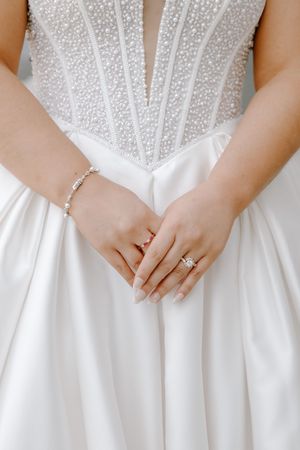 Close-up of bride's hands clasped together wearing a diamond engagement ring and pearl bracelet against a white satin wedding gown.