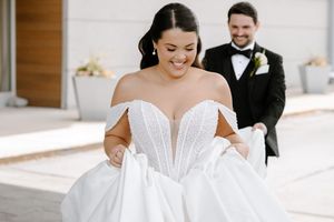 Bride in off-shoulder white ball gown smiles as groom in black tuxedo looks on during outdoor wedding.