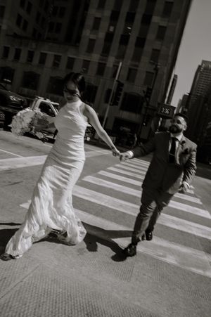 A bride in a white dress runs holding hands with a suited figure on a city crosswalk, captured in black and white.