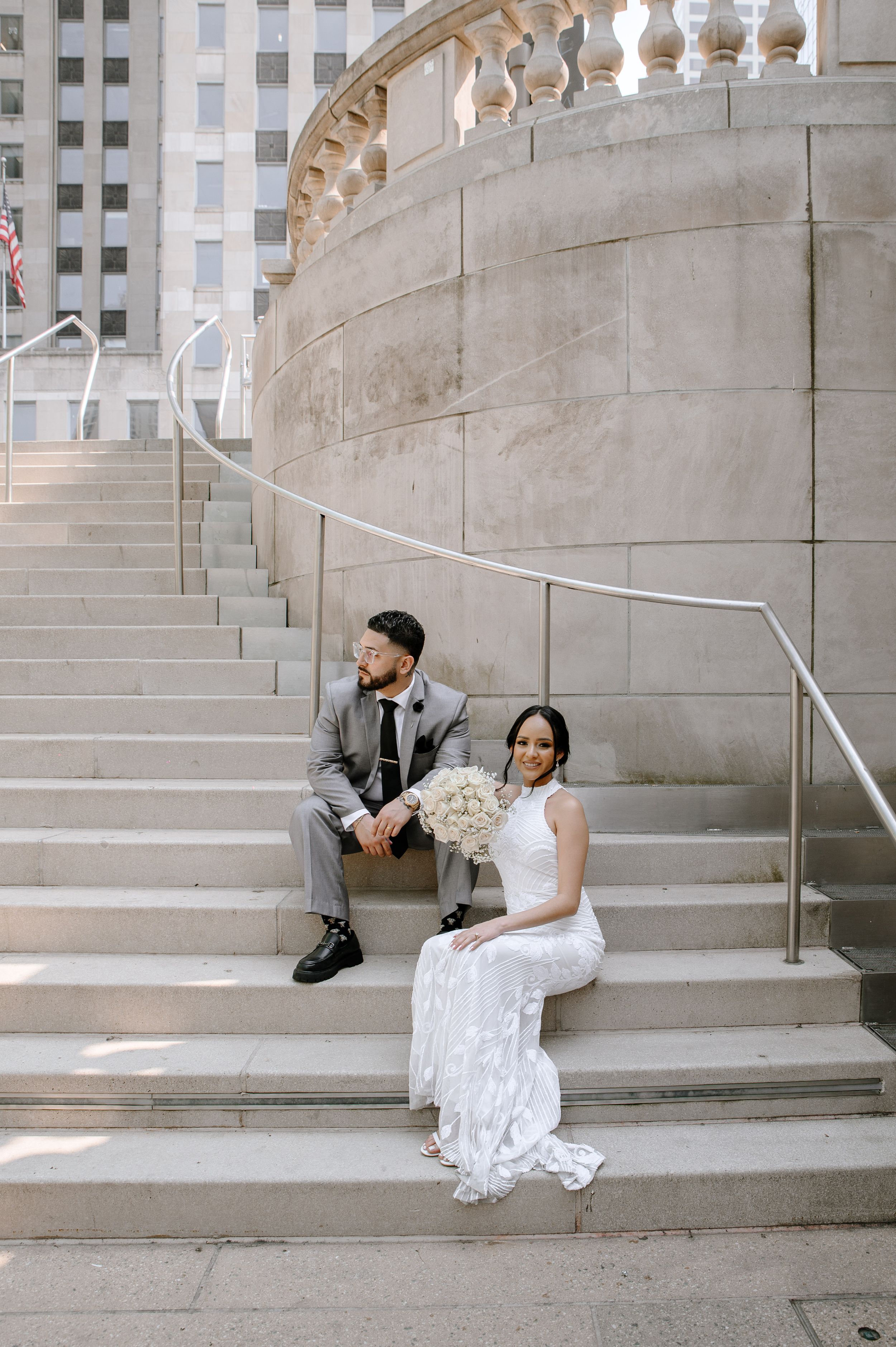 A couple in wedding attire sits on elegant stone steps with ornate balusters at an architectural venue.