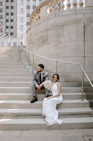 A couple in wedding attire sits on elegant stone steps with ornate balusters at an architectural venue.
