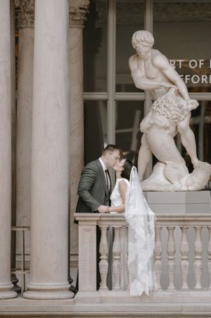 Bride and groom share a romantic kiss on a classical balcony with marble columns and elegant sculpture backdrop.