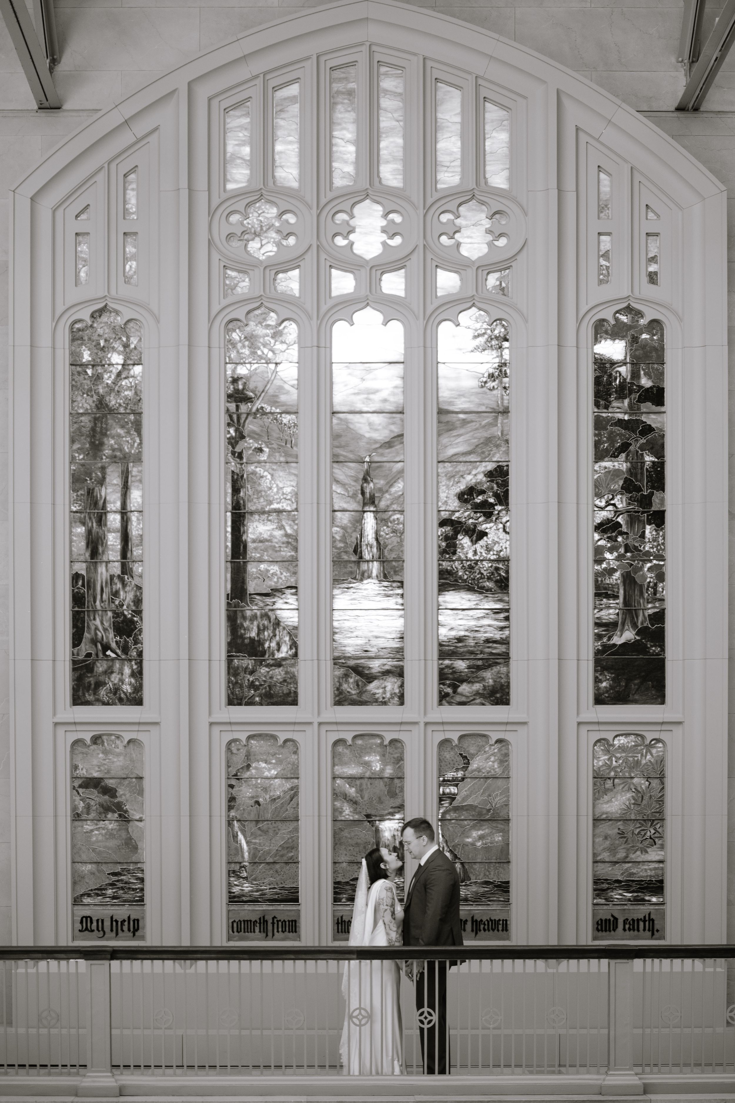 Bride and groom share a romantic moment before a stunning stained glass window in a historic church.