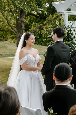 Bride in off-shoulder white gown and veil smiles at groom during outdoor wedding ceremony with green trees.