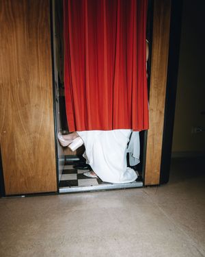 A person in a white dress and pink heels stands behind a red curtain in a wooden photo booth.