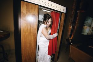 Bride in white wedding dress stepping into a vintage photo booth with red curtain at reception venue.
