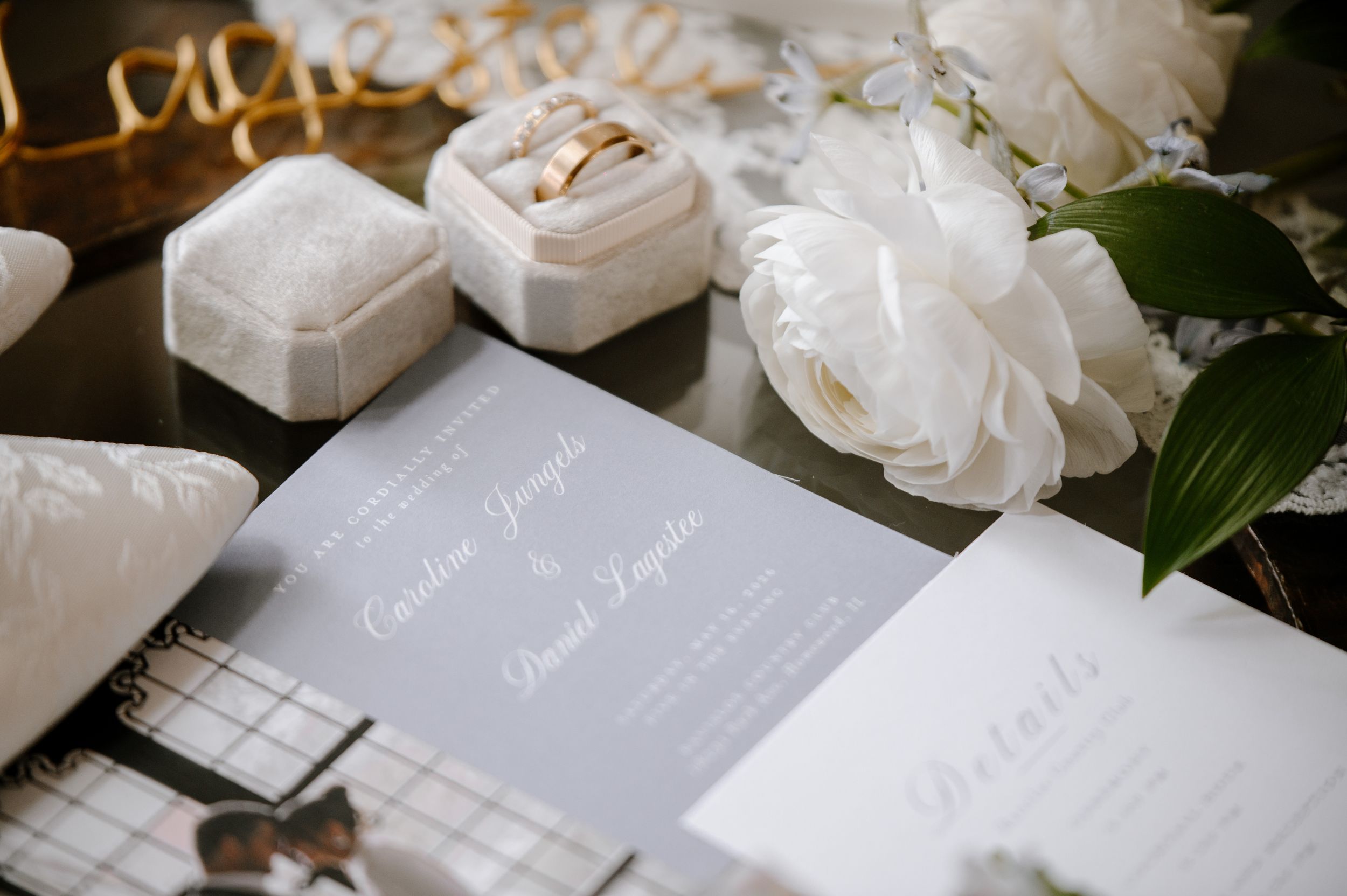 Elegant wedding flat lay with gray invitation suite, gold rings in velvet boxes, and white peonies on dark surface.