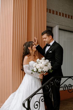 Bride and groom share a tender moment on a balcony, bride holding white rose bouquet in elegant off-shoulder gown.