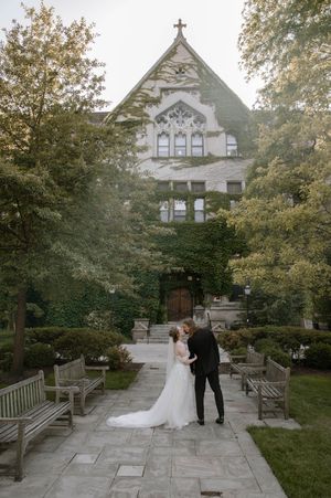 A couple in wedding attire stands on a stone path leading to a Gothic revival church surrounded by lush greenery.