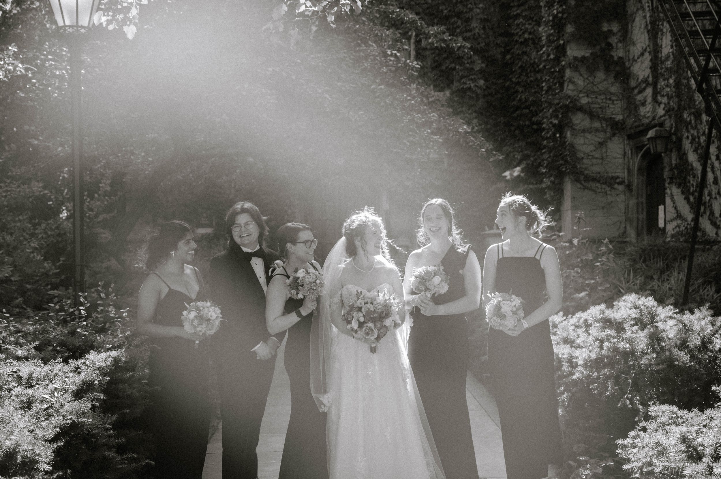 Wedding party members stand in a row on black and white film with flower bushes and trees in the background.