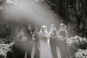 Wedding party members stand in a row on black and white film with flower bushes and trees in the background.