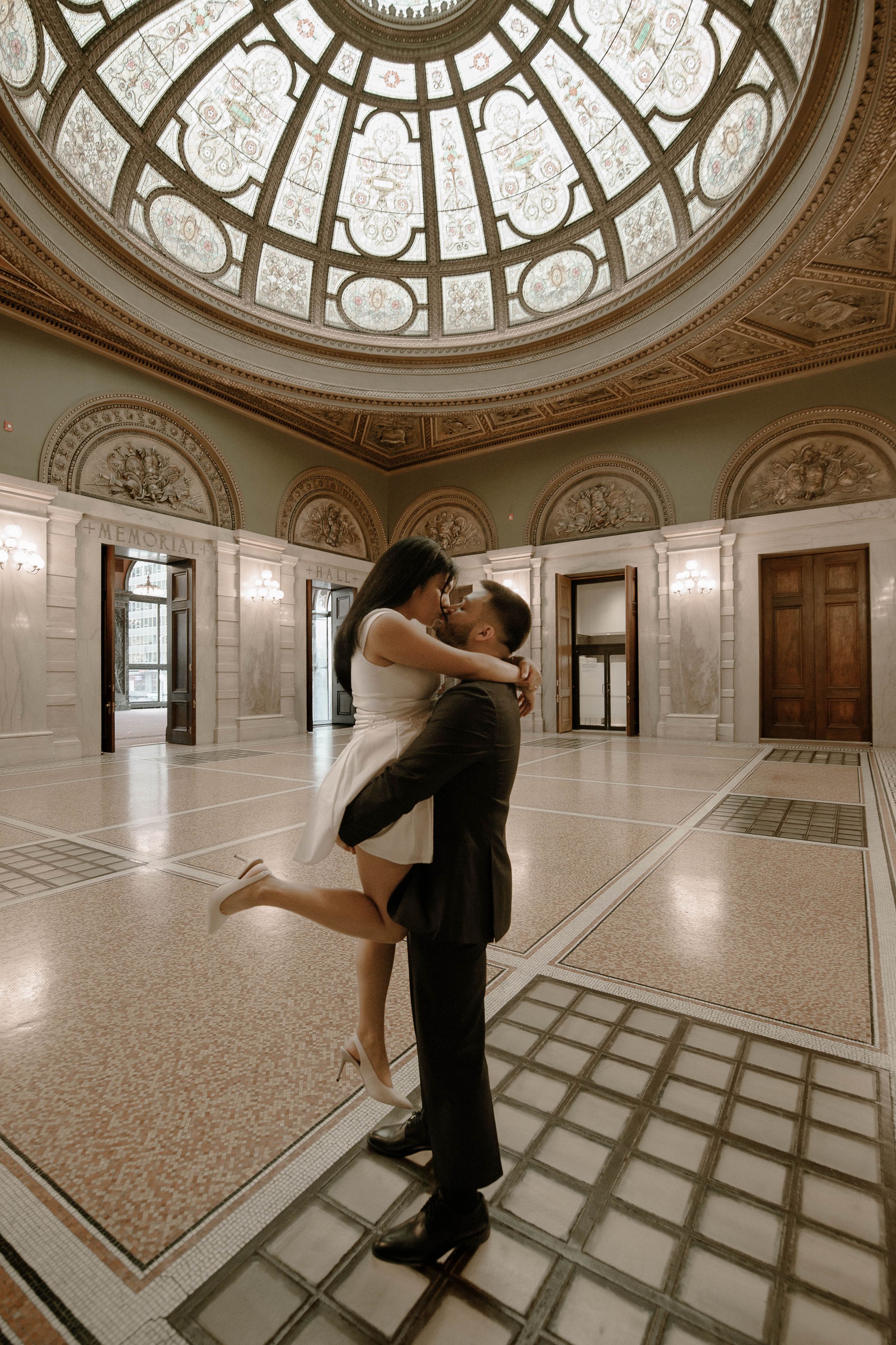 A romantic embrace in an elegant domed room with ornate neoclassical architecture and a grand skylight above.