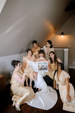 A group in cream-colored satin dresses gather on the floor looking at something together in an attic room with a lamp.