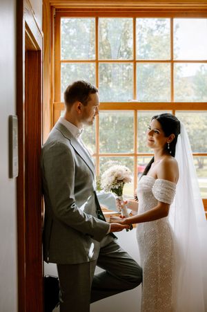 Romantic wedding couple shares intimate moments in a sunlit room with large windows and wooden trim.
