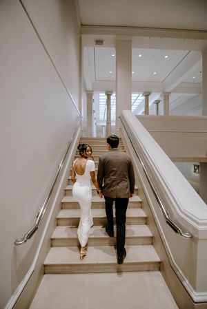 A couple walks up elegant white stairs holding hands, with the bride in a white dress and groom in a dark suit.