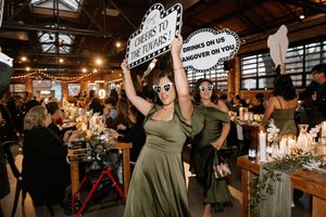 Bridesmaids in olive green dresses holding fun wedding signs at a reception.