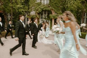 Wedding party walks outdoors past ivy-covered venue, bridesmaids in sage gowns and groomsmen in black tuxedos.