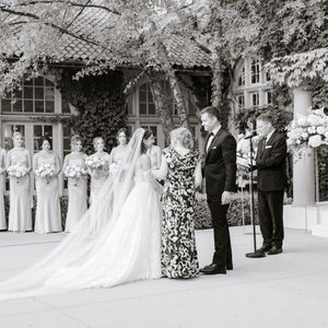 Black and white outdoor wedding ceremony with bride, groom, officiant, and bridesmaids beside a floral arch.