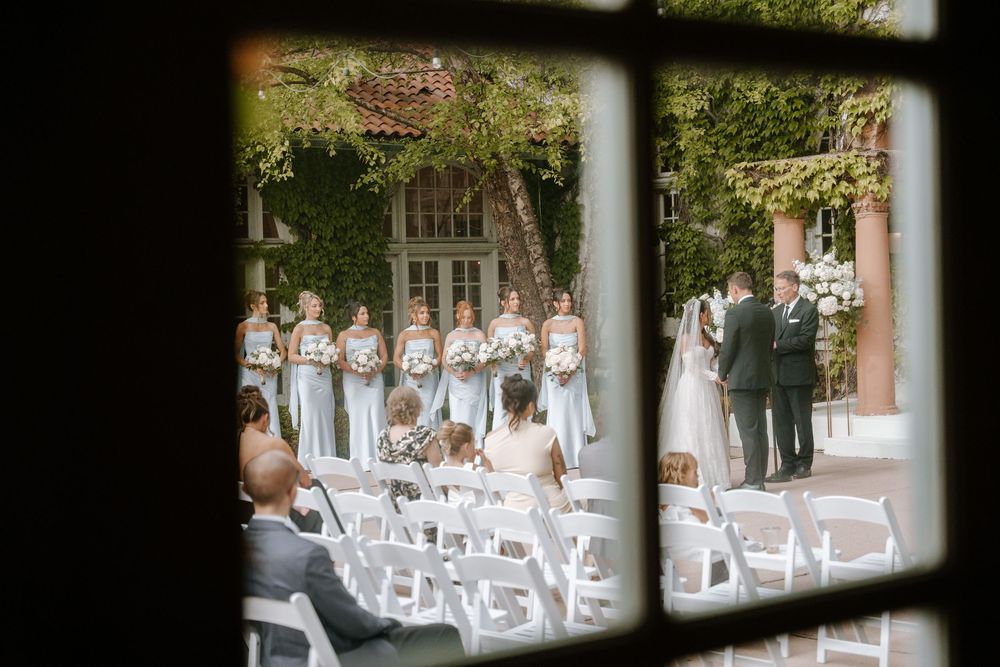 Outdoor wedding ceremony viewed through window, bride and groom at altar with bridesmaids in blue gowns at elegant venue.