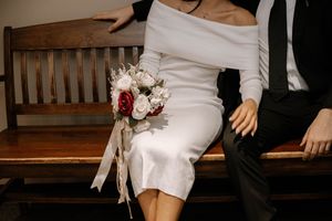 A bridal bouquet with white and burgundy flowers rests on a wooden bench next to a person wearing a white dress.
