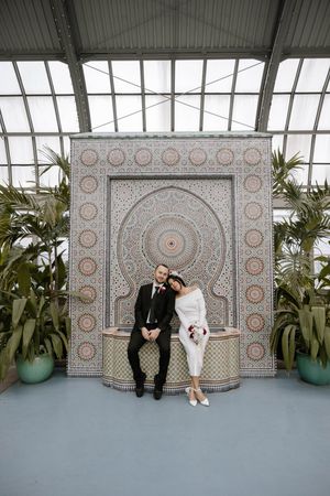 A couple poses in front of an ornate Moroccan-style tile wall backdrop with potted plants in an indoor greenhouse setting.