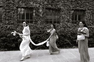 Black and white photo of a bride in white gown with two bridesmaids walking past an ivy-covered building.