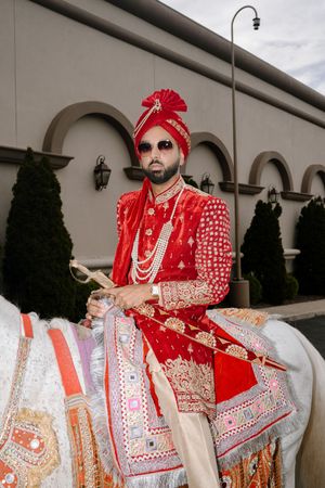 Groom in red sherwani and turban rides a white horse during traditional Indian baraat wedding procession.