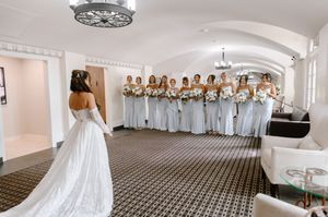 Bride in white gown faces her bridesmaids in silver dresses holding bouquets in an elegant venue.