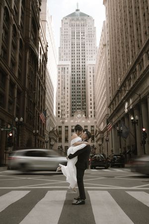 Groom lifts bride in wedding attire on a crosswalk in Chicago's financial district with tall buildings backdrop.