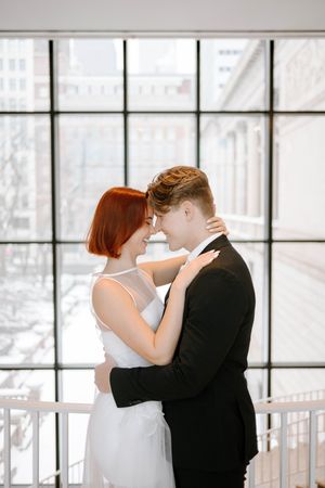 A romantic couple embraces by a window, with the person in white wearing a sleeveless dress and their partner in a black suit.