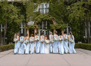 Bridesmaids in light blue gowns holding white bouquets stand together outside an ivy-covered venue.