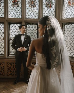Bride in white gown and lace veil faces groom in black tuxedo before stained glass windows in elegant venue.
