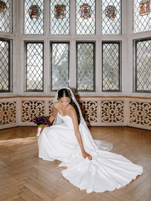 Bride in white gown and veil sits gracefully on herringbone floor before ornate latticed windows in elegant venue.