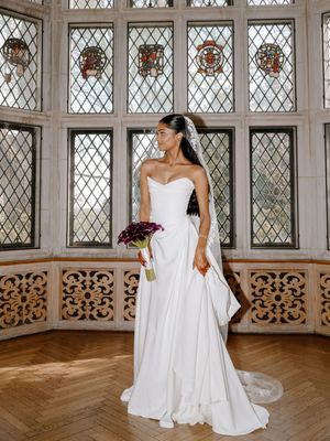 Bride in strapless white gown holding purple bouquet stands before ornate stained glass windows in elegant chapel.
